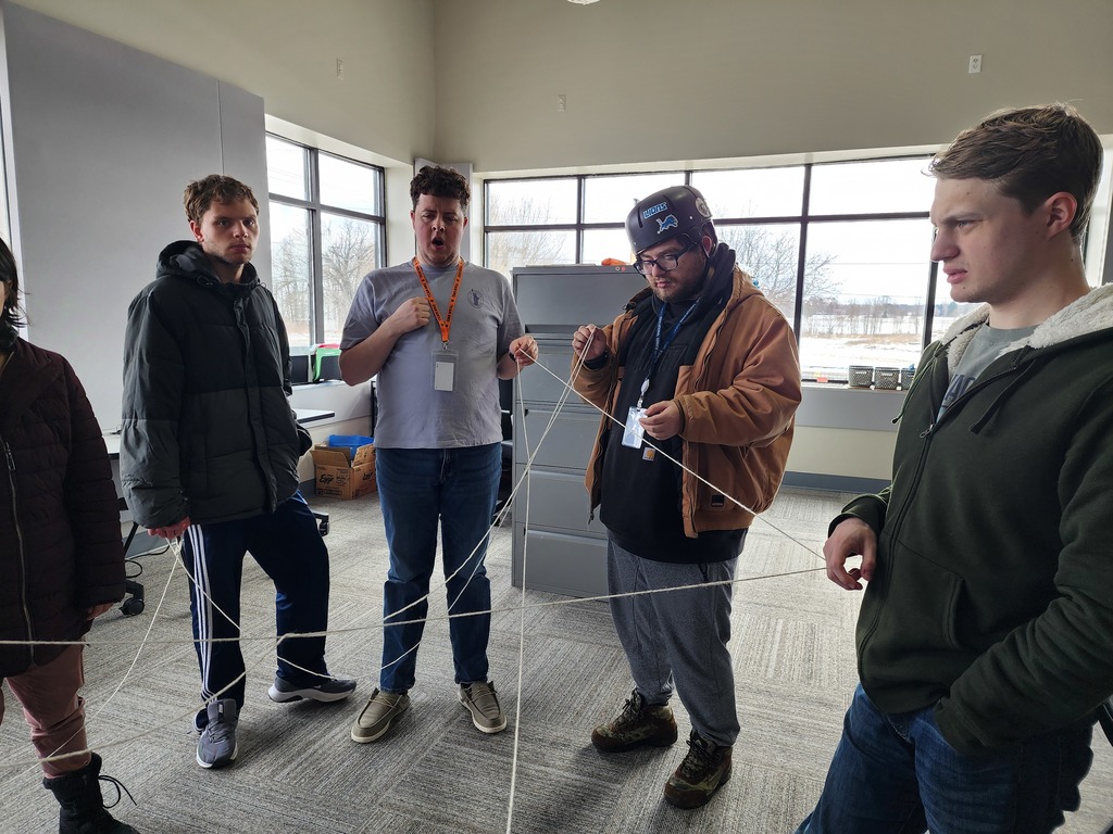 Young adult males standing in part of a circle with string hanging from their fingers.