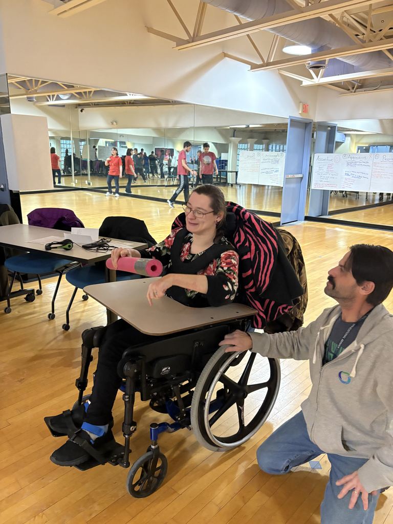  A young female adult in a wheel chair playing an adaptive game while an adult male assists.