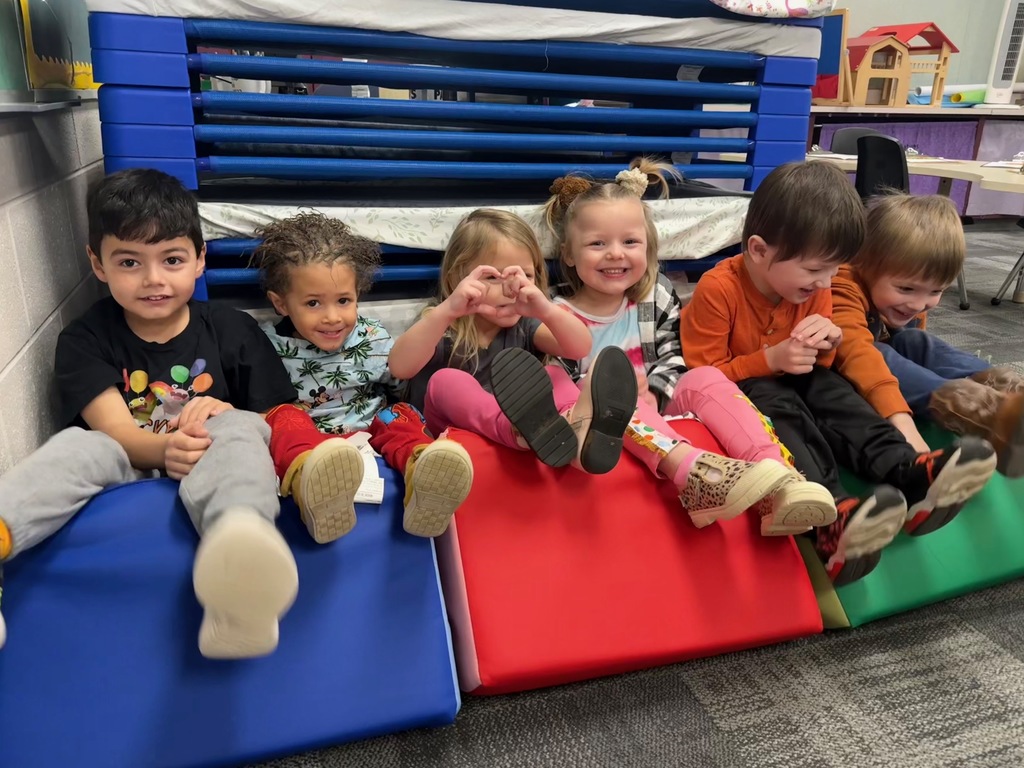 Six preschool children sit together on upside-down colorful cushions in a classroom during Wacky Wednesday, smiling and laughing together.