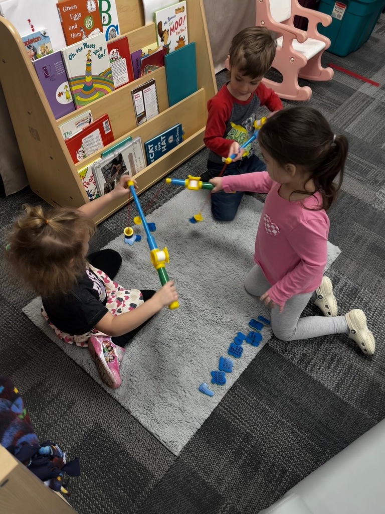 Preschool children sit on a rug using toy fishing rods to catch number pieces and count the raised dots on the back.