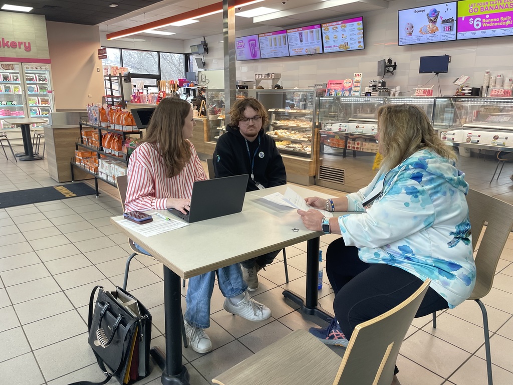 Three people sitting at a table inside a Dunkin’ restaurant, reviewing documents and looking at a laptop.