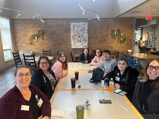 A group of students and staff members sitting around a table together in a common area, smiling for a photo.