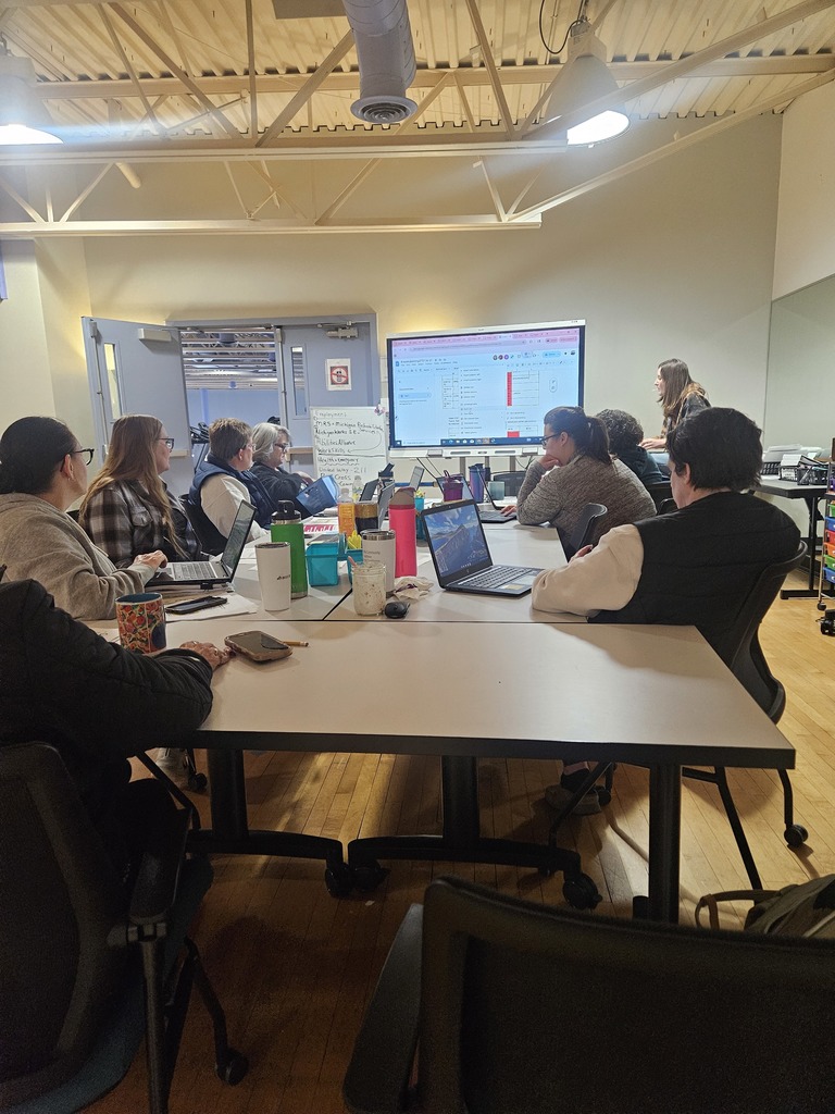 The photo shows a group of about 8 to 10 adults engaged in a professional meeting or workshop. They are seated around several large, light-colored rectangular tables that have been pushed together to form a long communal workspace.