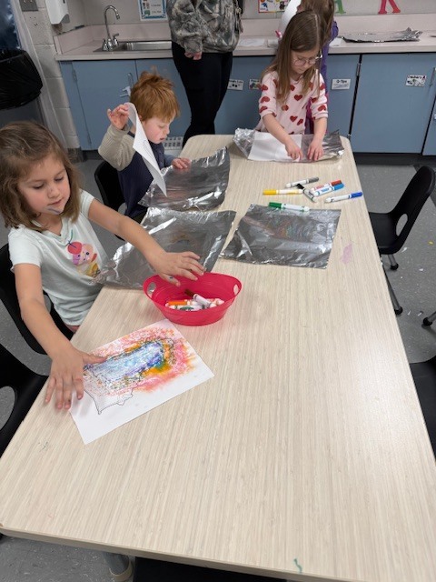 Preschool children coloring on foil sheets with markers, spraying them with water, and pressing paper to transfer the colors at a classroom table.