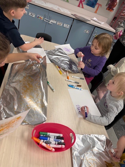 Preschool children coloring on foil sheets with markers, spraying them with water, and pressing paper to transfer the colors at a classroom table.