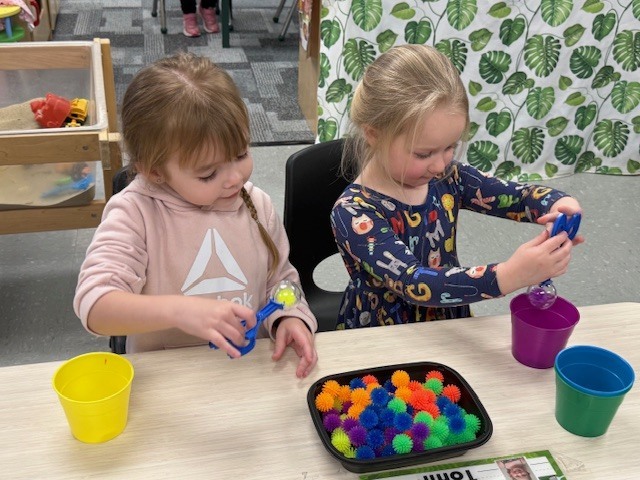 Two preschool children use scoops to pick up colorful pom poms and place them into cups, sorting them by color at a classroom table.