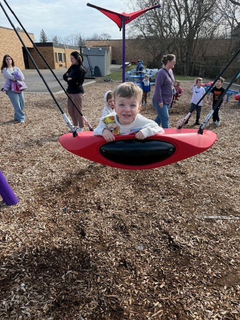 A preschool child smiles while lying on a red playground swing as adults and children stand and play in the background.