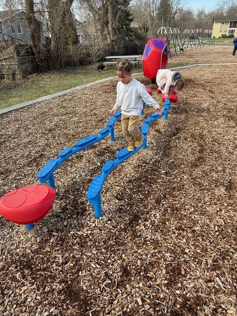 Preschool children balance and walk across blue playground equipment while playing outside on a woodchip-covered surface.
