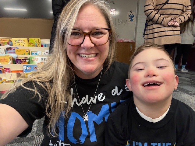 Teacher and young student smiling together in a classroom with books behind them.