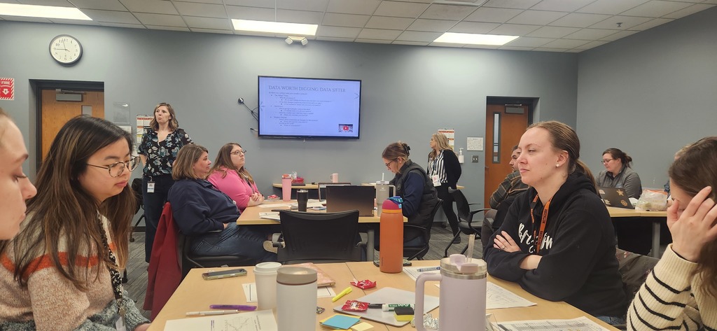 A woman talking at the front of a room with a presentation on a TV. Three groups of tables with people sitting at all of the tables can be seen.