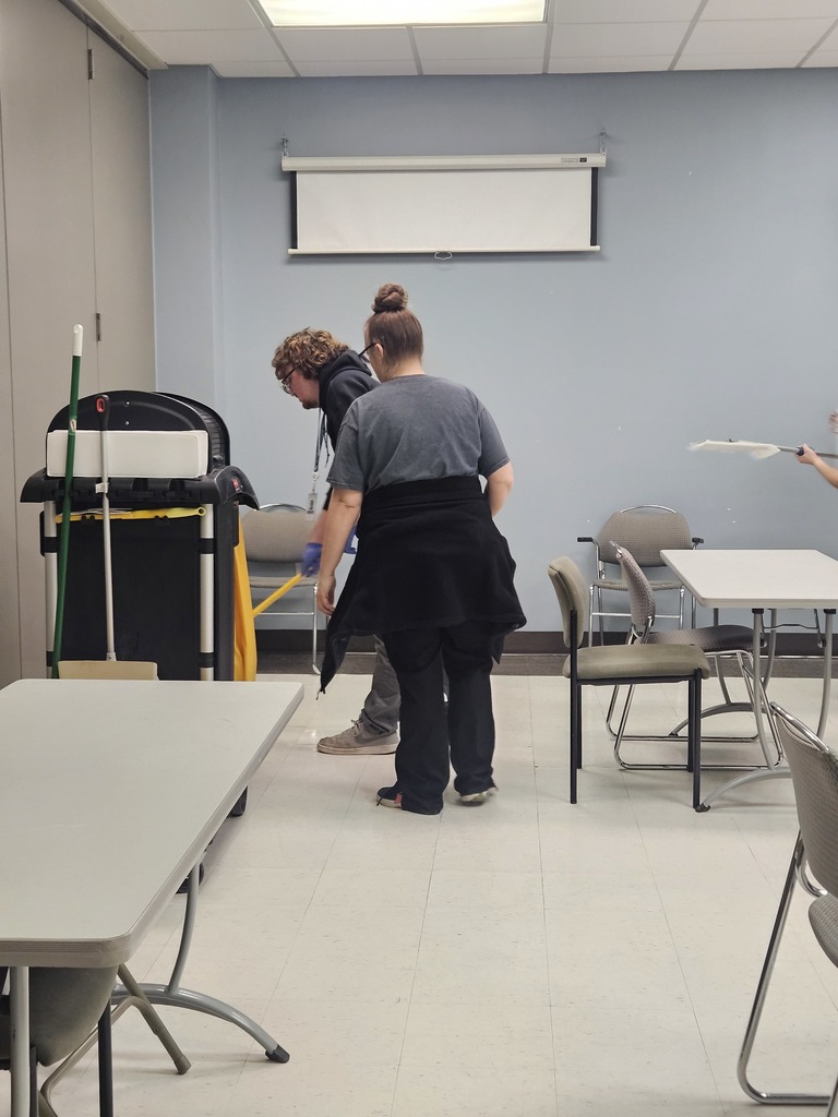 Two people working near a large professional cleaning cart in a classroom or meeting space
