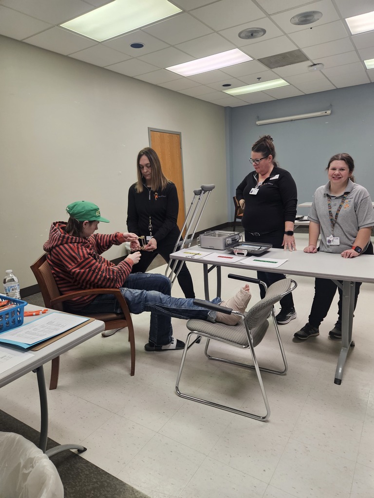A group in a medical training setting with one person's leg propped up on a chair with a cast