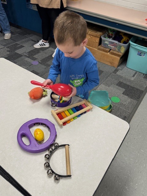 A preschooler sits at a table holding a toy instrument while exploring various percussion instruments like a tambourine, xylophone, and maracas.