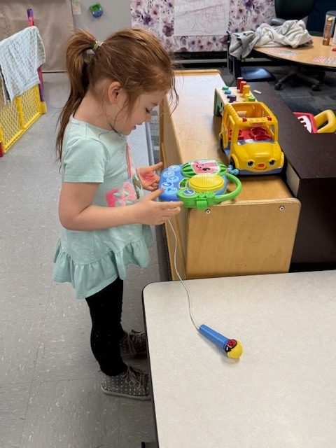 A preschooler stands near a table, smiling and engaged as she listens to music playing from a speaker.