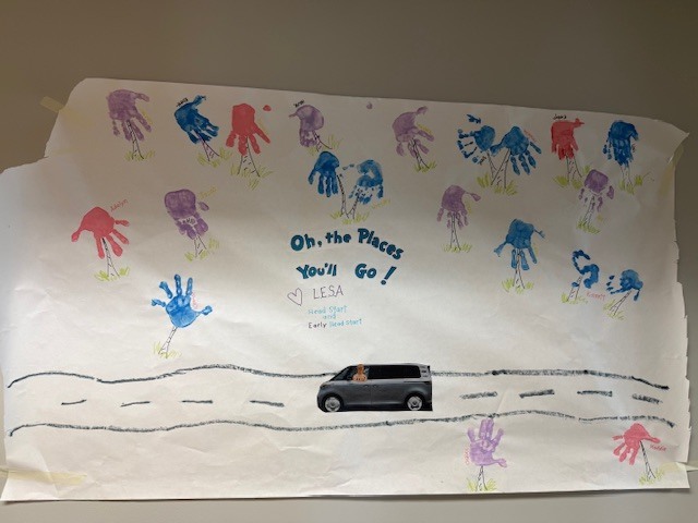 A decorated poster with colorful handprints and a road scene celebrates an early learning leader during a clap-out, with a car driving forward and the message “Oh, the Places You’ll Go!” displayed.