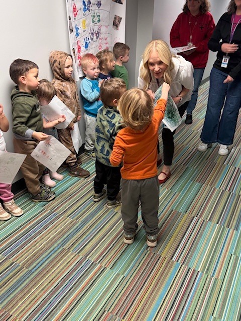 Preschool children stand in a hallway holding papers while two children hand their papers to a crouching staff member, who smiles as she receives them..