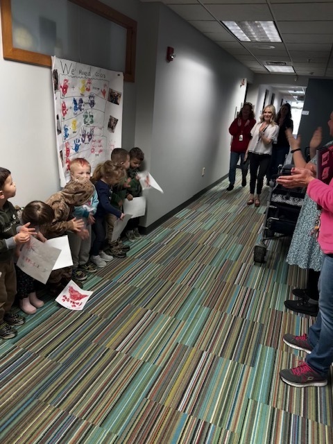 A group of preschoolers line the wall holding artwork, smiling and clapping as a staff member walks down the hallway with colleagues behind her.