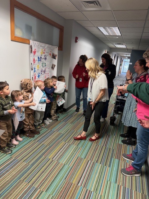 Preschool children stand in a hallway holding papers while adults clap as a staff member walks up to them.