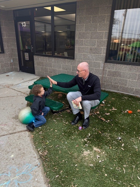 Superintendent sitting at an outdoor table giving a high five to a child sitting on a large bouncing ball.