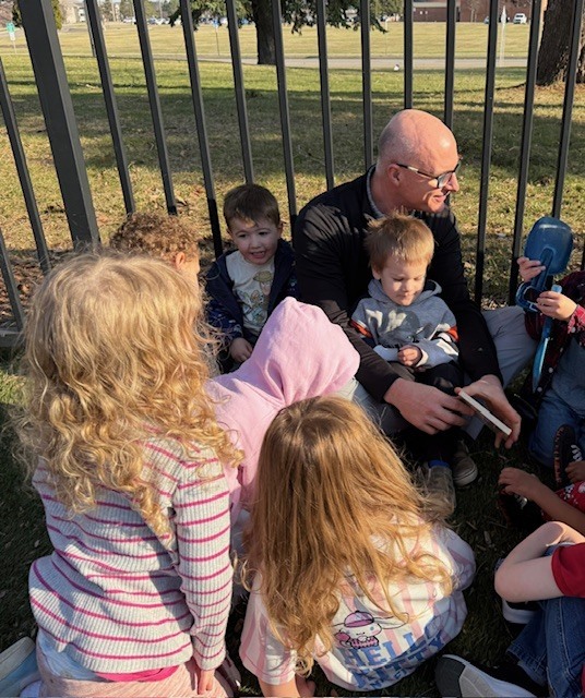 A man sitting on the grass reading a book to a group of young kids.