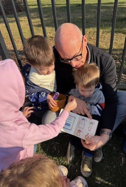 A young kid sitting in the lap of a man with glasses, reading a book, surrounded by other young kids listening intently.