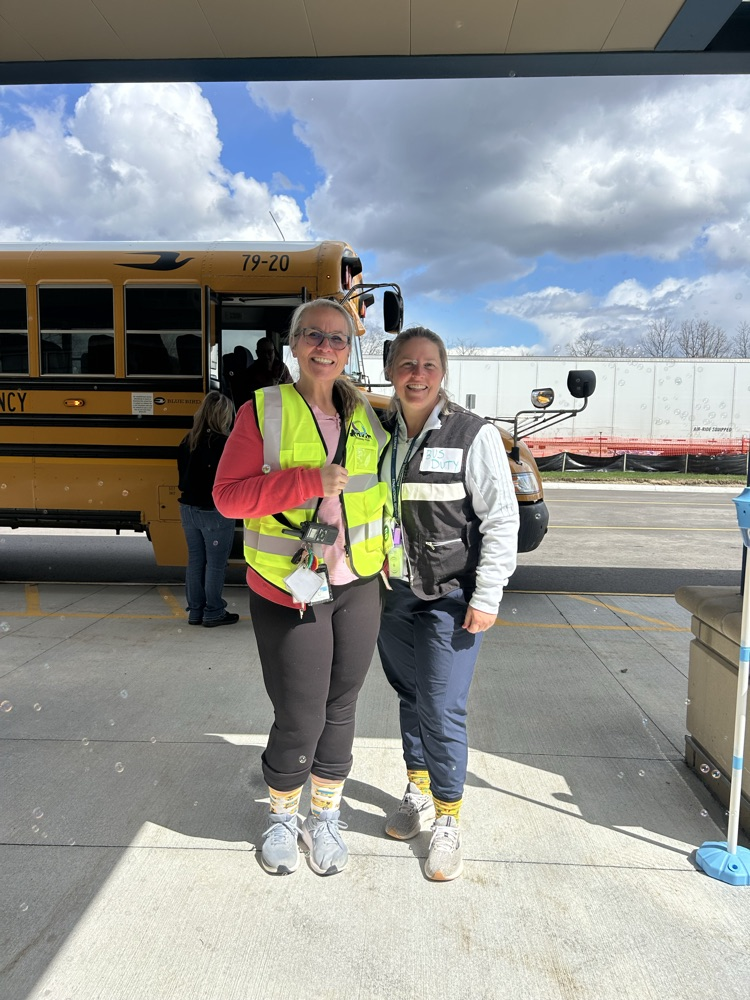 Two woman standing together, arm in arm, smiling in front a bus.