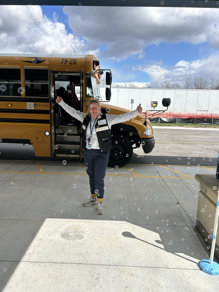 A woman smiling with her thumbs up, standing in a bus lane. A bus is in the background and bubbles are floating in front of her.