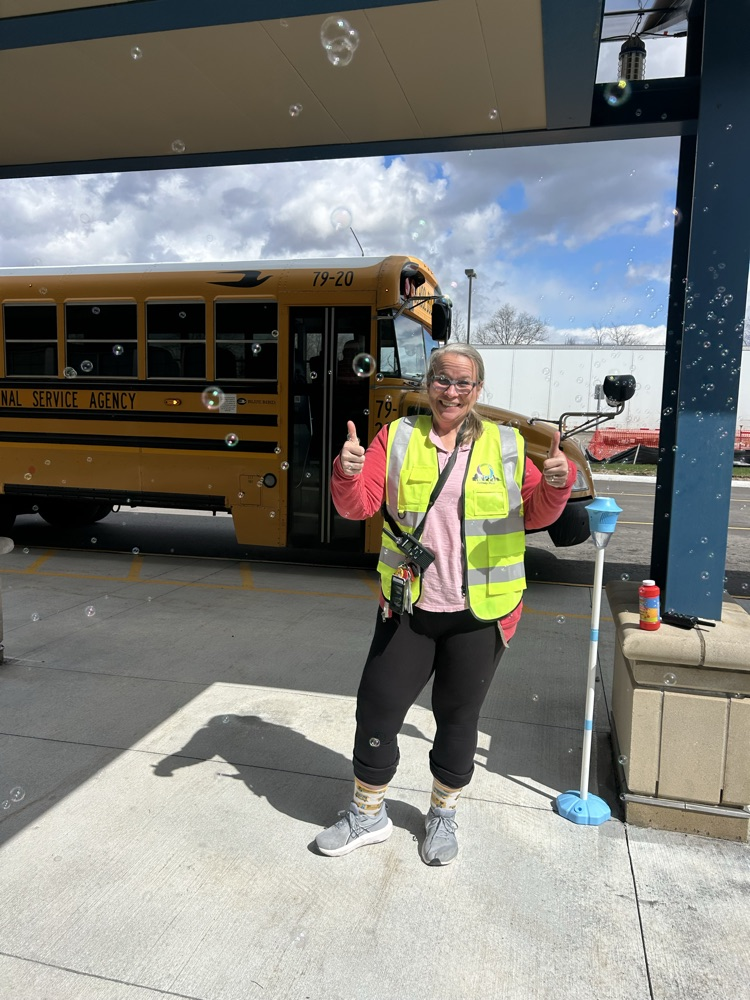 A woman in a bright, reflective vest, smiling with her thumbs up, standing in a bus lane. A bus is in the background and bubbles are floating in front of her.