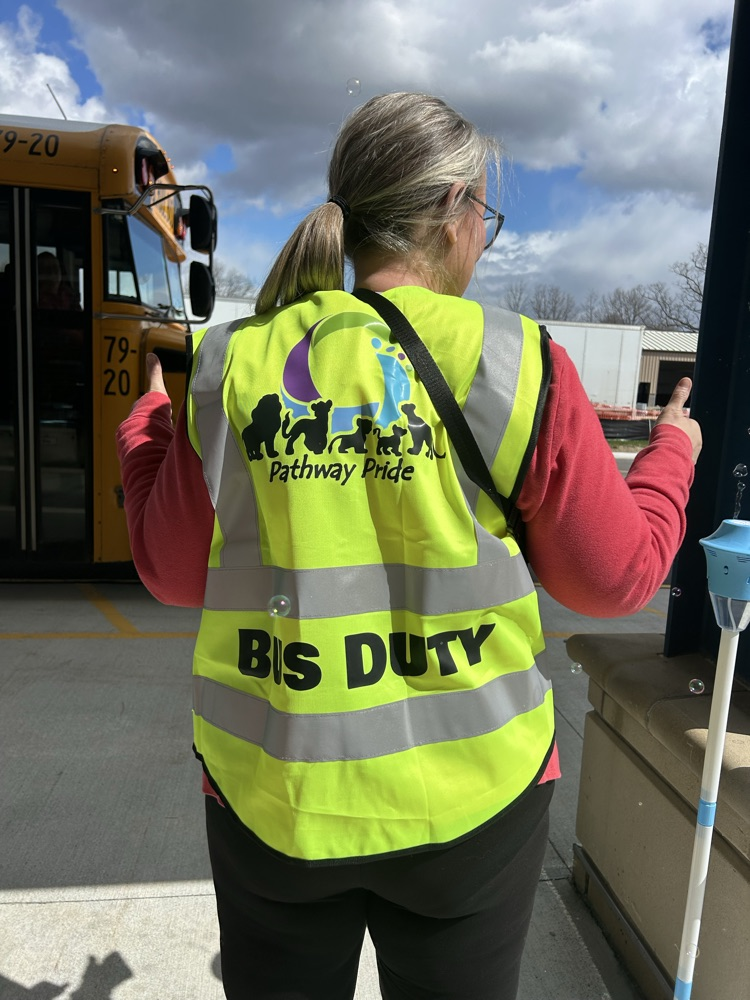 A woman with her hair pulled back is standing with her thumbs up. Her reflective vest says "Pathway Pride" and "Bus Duty".