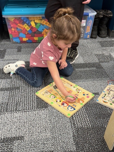 Preschool child kneeling on classroom carpet, using a wooden board to sort colorful pieces by color.