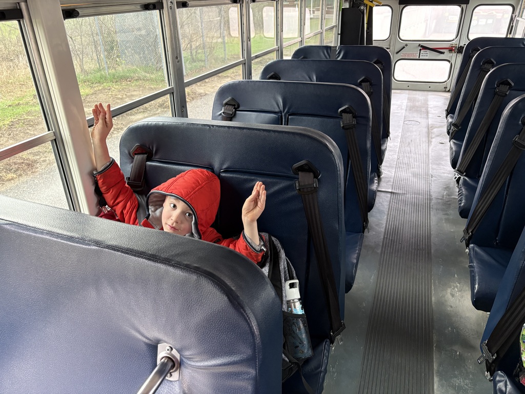 A young boy in a red coat is sitting on a bus. He has his hands up doing 'bunny ears" at a railroad stop.