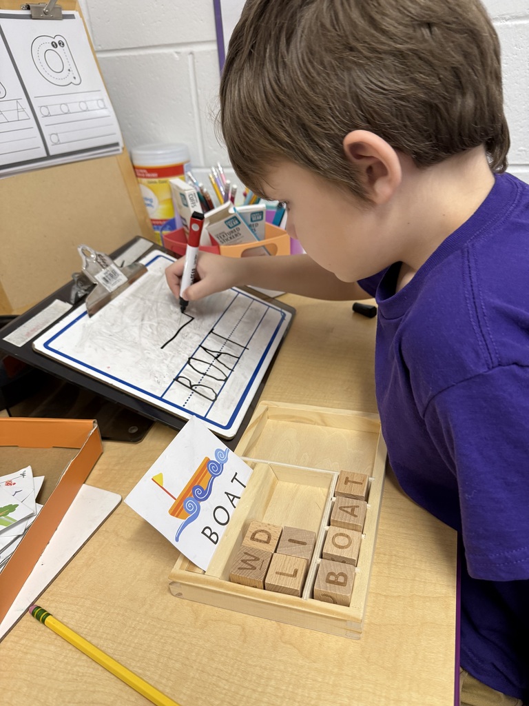 A young boy with brown hair sits at a desk, focused on a spelling activity. He is placing a wooden block with the letter "T" into a tray to complete the word "BOAT," which matches an illustrated card nearby. In the background, a small whiteboard displays the word "BOAT" written in large, clear letters.