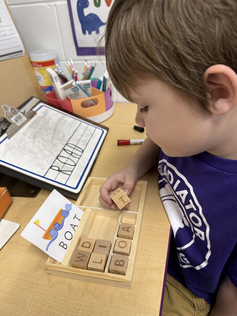 A young boy with brown hair sits at a desk, focused on a spelling activity. He is placing a wooden block with the letter "T" into a tray to complete the word "BOAT," which matches an illustrated card nearby. In the background, a small whiteboard displays the word "ROAD" written in large, clear letters.