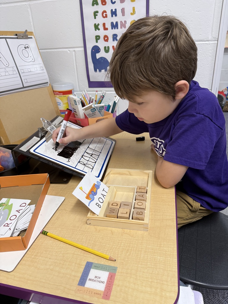 A young boy with brown hair sits at a desk, focused on a spelling activity. He is placing a wooden block with the letter "T" into a tray to complete the word "BOAT," which matches an illustrated card nearby. In the background, a small whiteboard displays the word "BOAT" written in large, clear letters with a drawing of a boat drawn by the boy.