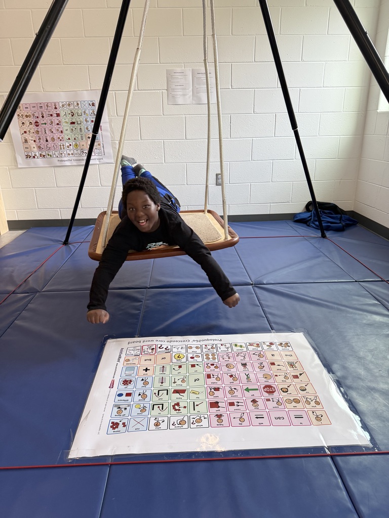 A young person with a joyful expression lies on their stomach on a flat platform swing, suspended from a black metal frame. They are reaching their arms forward toward a large, laminated communication board (AAC) laid out on blue mats on the floor. The board features a grid of colorful symbols and words used for non-verbal communication. In the background, white cinderblock walls feature another communication poster and a few papers.
