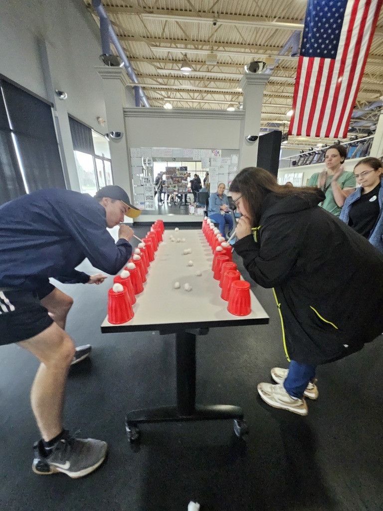A wide-angle, eye-level shot captures two young people engaged in a competitive "Minute to Win It" style game on a long white table in a gym. A young man on the left, wearing a blue jacket and black shorts, and a young woman on the right, in a black jacket and jeans, lean over the table. Both use straws to blow white cotton balls off the tops of several red plastic cups lined up along the edges of the table. More cotton balls are scattered across the center of the table. A few spectators watch from the background, and an American flag hangs on the wall to the right.