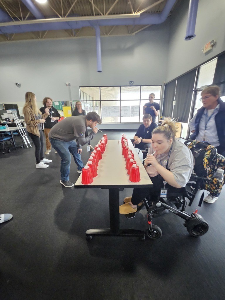 A wide-angle, eye-level shot captures a group of people in a large, modern indoor space with high ceilings and purple structural pipes. In the center, a man and a woman in a motorized wheelchair are competing in a game at a long white table. Both lean forward, using straws to blow white cotton balls off the tops of two parallel rows of red plastic cups.  Several onlookers stand nearby, watching the competition with smiles. Large windows in the background reveal a bright hallway, and the room features dark flooring and industrial-style lighting.