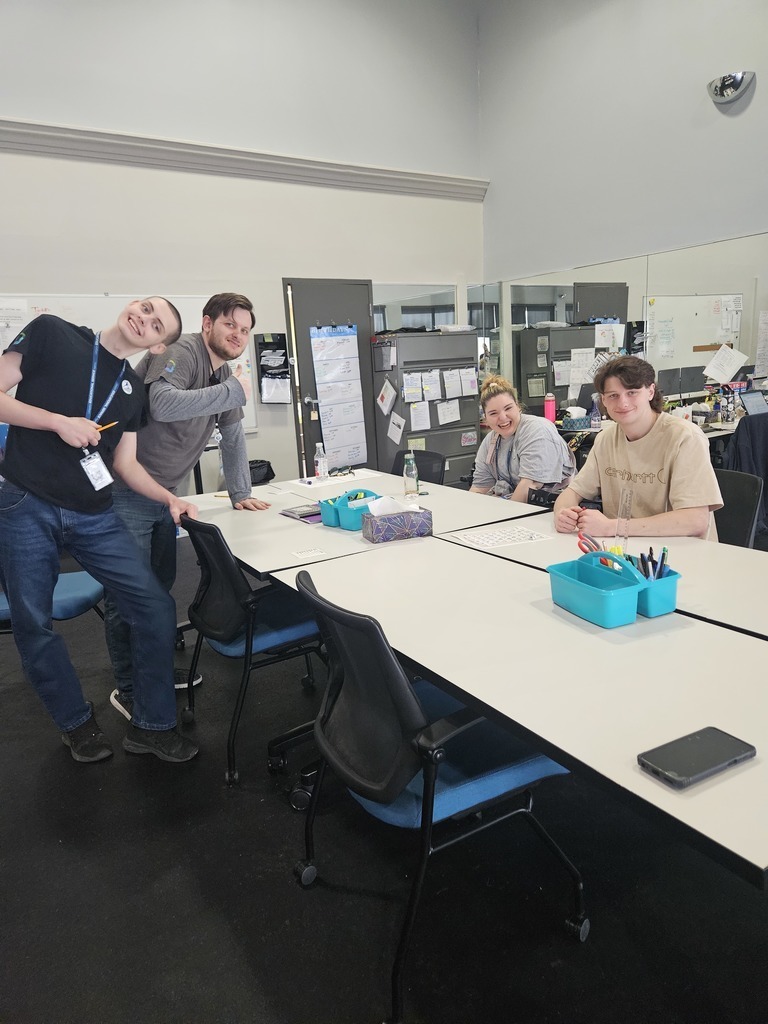 A bright, medium shot shows four young adults gathered around a large white conference table in an office or classroom setting. Two young men stand on the left, one leaning in with a smile and the other giving a thumbs-up. To the right, a young woman and another young man are seated, looking toward the camera with friendly expressions. The table is neatly organized with turquoise supply caddies and tissues. In the background, there is a large wall mirror, filing cabinets, and a door with a calendar, creating a functional and collaborative atmosphere.