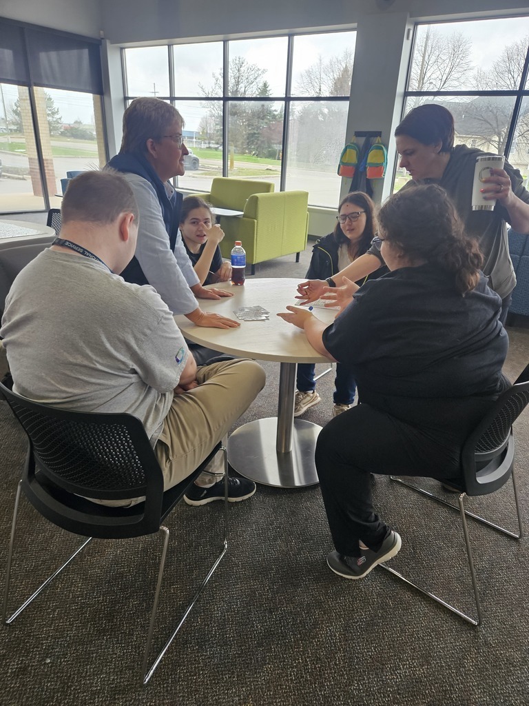 A high-angle, medium shot shows a group of six people gathered around a small, round white table in a bright common area. Two people are seated in black chairs in the foreground, while others stand or sit around the table, engaged in a lively discussion or game. Large floor-to-ceiling windows in the background fill the room with natural light and overlook a parking lot and green space. The setting is casual and communal, featuring modern furniture like a lime green armchair and carpeted flooring.