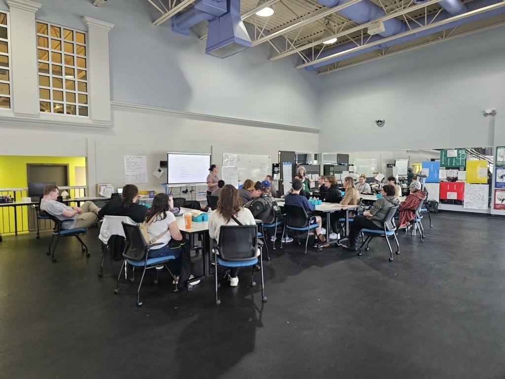 A wide-angle, eye-level shot shows a group of about fifteen young adults seated at clustered tables in a large, open industrial-style room. They are facing a woman standing at the front by a digital whiteboard, appearing to lead a presentation or class. The room features high ceilings with exposed blue pipes, dark flooring, and a large mirror along the right wall that reflects the space. The setting is bright and functional, suggesting a collaborative workshop or educational environment.