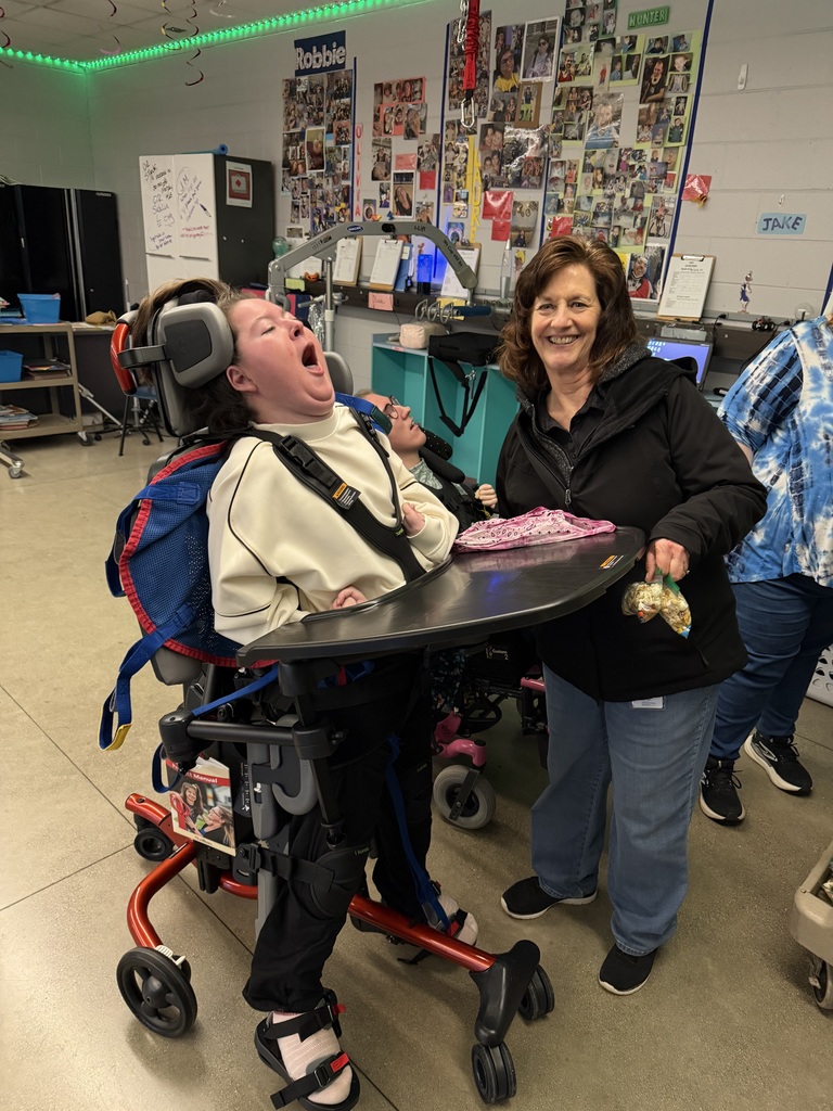 A student sits in a supportive chair with a tray, wearing headphones. The student is in a classroom with colorful photo boards on the wall. A smiling adult stands next to them holding a bag of trail mix. Another student and adult are nearby in the background. Staff visitors are present, showing support and encouragement.
