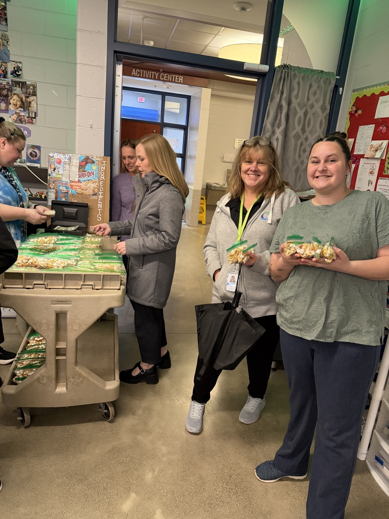 A group of adults stands in a school hallway near a cart filled with small bags of trail mix. Two women in the front smile and hold up the snack bags. Other staff members are helping organize and sell the treats. A display shows the “pup” themed trail mix. Staff from another part of the building have come to support the students.