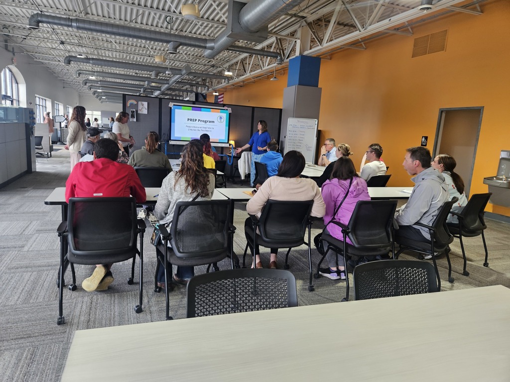 A group of people sitting at tables in a large, open office space watching a presentation on the PREP Program.