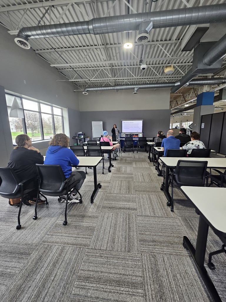 A classroom setting where several people sit at tables and listen to a presentation on a screen at the front of the room.