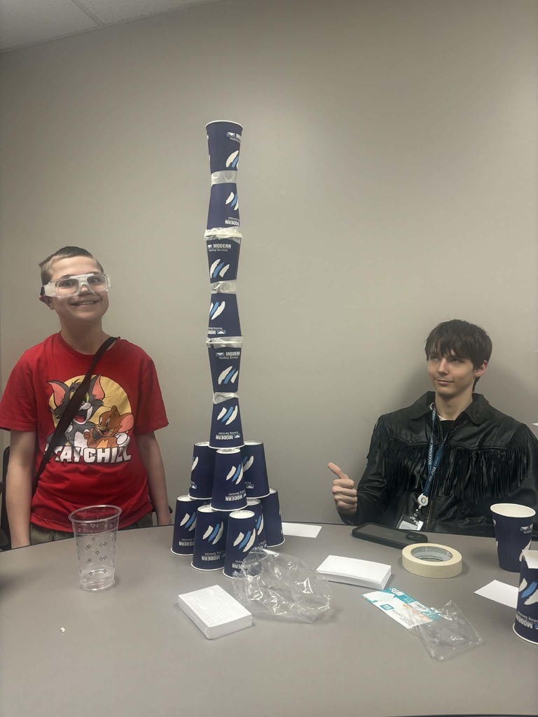 Two students smiling in front of a tower of cups stacked on a table. Cups, tapes, and notecards are scattered on the table below.