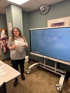 A smiling woman stands next to a large digital whiteboard that has the words "Happy Internship Day!" written on it
