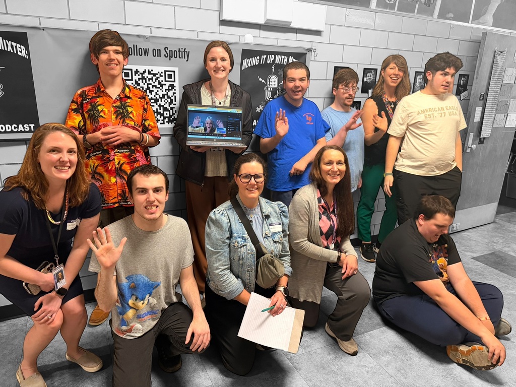 A group of ten people, including students and adults, pose for a photo in a room with light-colored brick walls. They are gathered in front of posters, one of which features a large QR code and the text "Follow on Spotify." One woman in the back row holds up a laptop displaying a person on a video call. A young man in the front row wears a grey shirt featuring Sonic the Hedgehog and waves at the camera, while another young man sits on the floor to the right. The group appears to be in a classroom or media space, smiling and engaged.