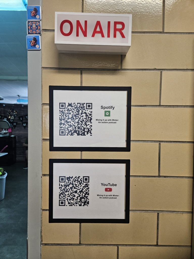 A close-up shot of a tan tiled wall featuring a white "ON AIR" sign with bold red lettering at the top. Below the sign, two framed papers are mounted vertically, each displaying a large QR code. The top paper includes the Spotify logo and the bottom includes the YouTube logo, both accompanied by the text "Mixing it up with Mixter: An autism podcast." To the left, several small podcast-themed stickers are visible on the edge of the wall.
