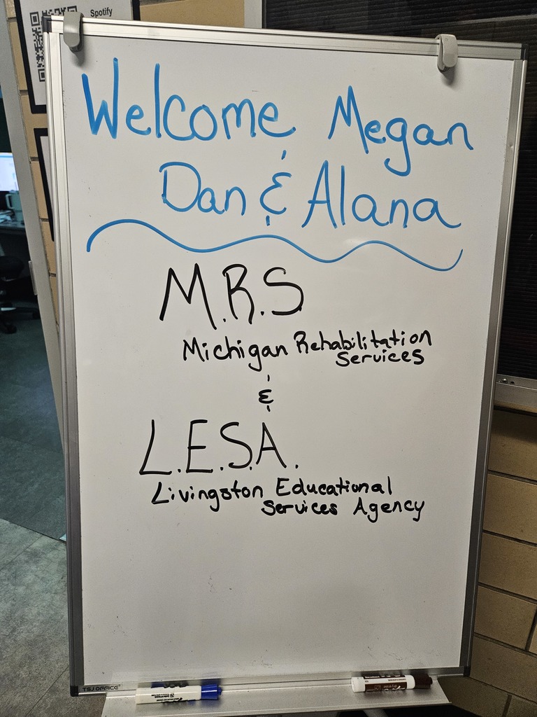 A white mobile whiteboard features handwritten text in blue and black markers. At the top, in blue, it says "Welcome Megan, Dan & Alana" above a wavy blue line. Below the line, in black, it lists "M.R.S. Michigan Rehabilitation Services" and "L.E.S.A. Livingston Educational Services Agency." Dry-erase markers rest in the tray at the bottom of the board.
