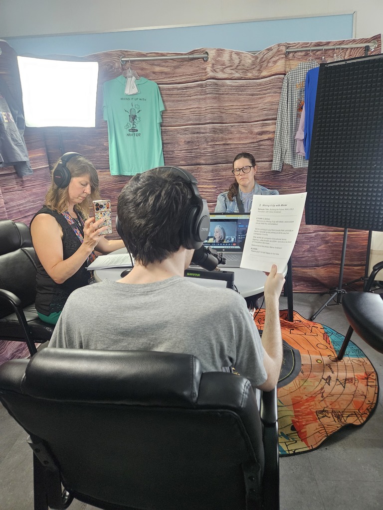 Three people are in a small podcast studio with a wood-paneled backdrop. In the foreground, a person with headphones sits with their back to the camera, holding a script. Across the table, a woman with glasses listens, while another woman to the left wears headphones and holds up a phone. A laptop on the table shows a person on a video call, and professional lighting and sound equipment are visible around the group.